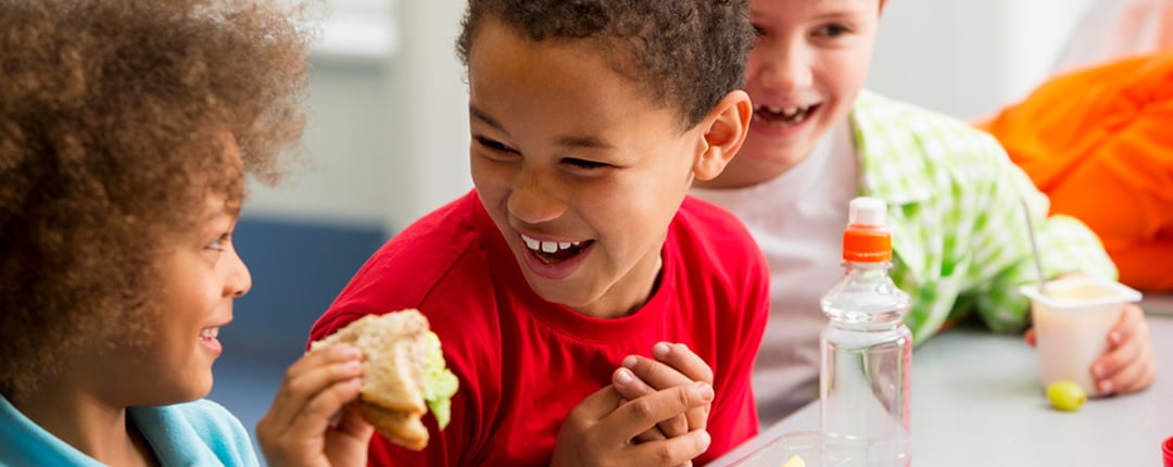 A group of three kids share a friendly moment during lunch, surrounded by healthy foods including fruit cups, yogurt, and a sandwich.