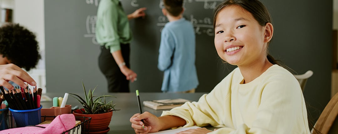 Student seated at a classroom table writing in a notebook, with pencils and school supplies nearby, while an instructor and another student work at a chalkboard in the background.