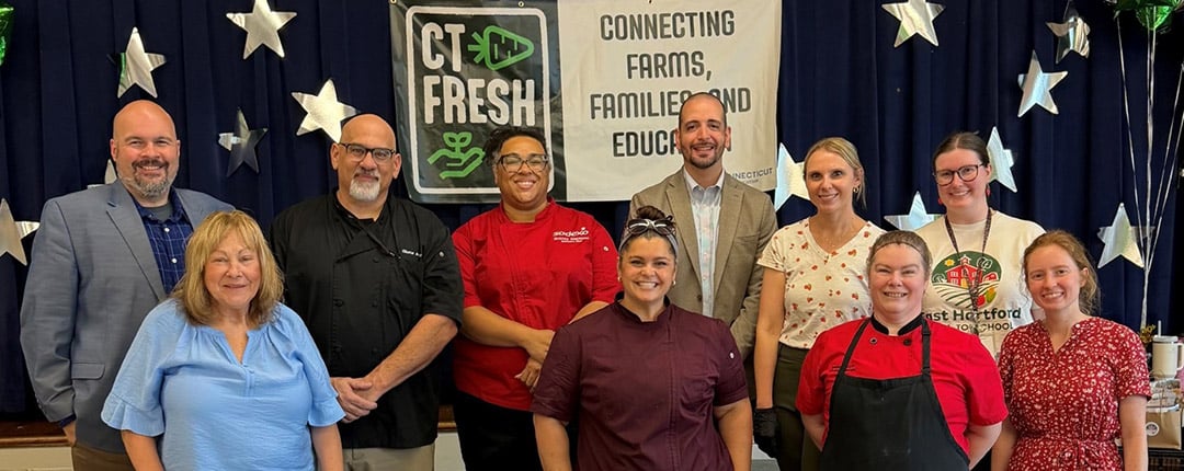 School nutrition professionals, educators, and community partners gathered around a table displaying fresh strawberries and prepared food samples at a farm-to-school event promoting local food and student wellness.