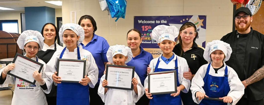 Children in chef uniforms holding achievement certificates alongside culinary mentors during a school cooking competition, highlighting student learning, teamwork, food education, and culinary skills development.