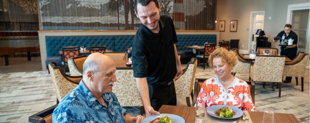 A dining staff member serves food to a resident couple in a senior living community dining room.