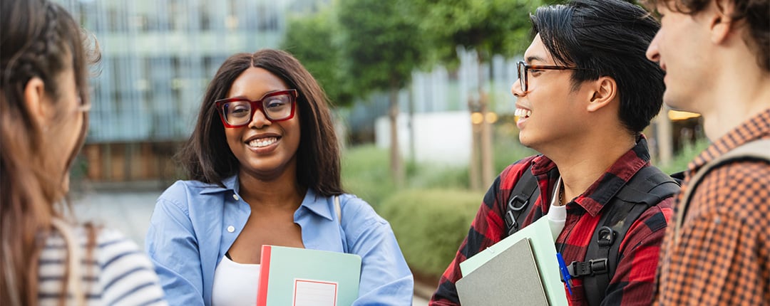 Students standing outdoors on a campus walkway, holding notebooks and backpacks while talking together near modern buildings and green landscaping.