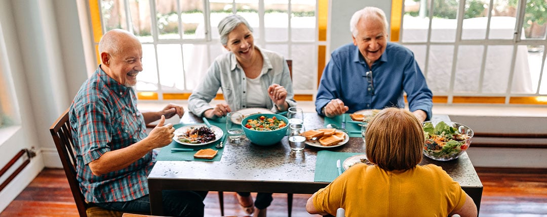 Older adults gathered around a dining table sharing a meal with salads, vegetables, and refreshments in a bright community setting, highlighting social connection, healthy dining, and wellness-focused living.
