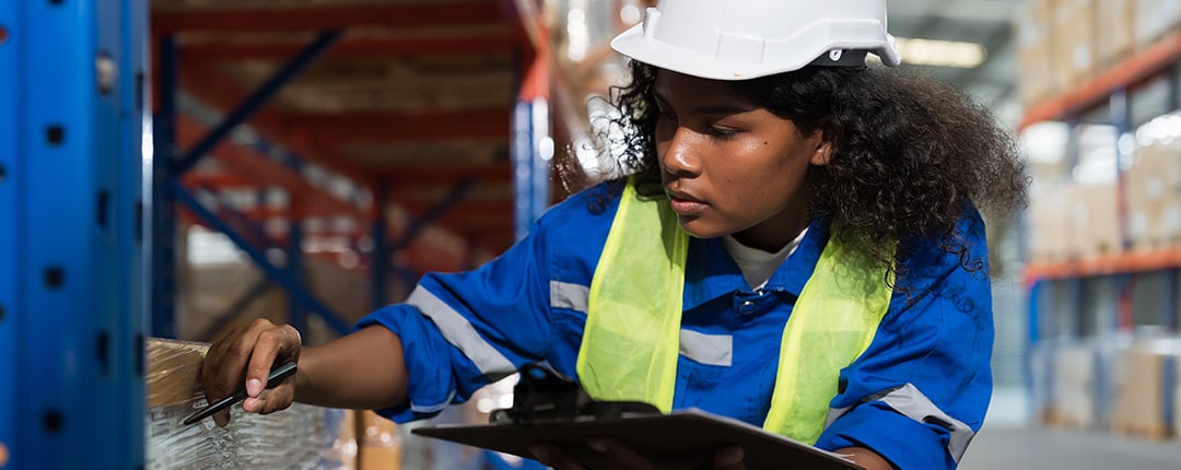 Warehouse worker in safety gear inspecting packaged inventory with a clipboard in a distribution center, highlighting facility management, inventory control, logistics, and operational efficiency.