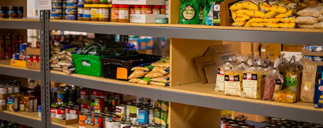 Shelves stocked with packaged foods including rice, pasta, canned goods, and jars, arranged in a storage or pantry area with labeled boxes and organized sections.