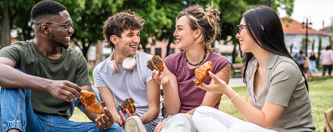 Four people sitting on a picnic blanket in a park, holding pastries and talking together. 