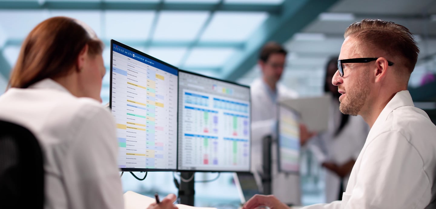 Healthcare professionals working at a computer workstation with dual monitors displaying schedules and data dashboards, while colleagues collaborate in the background of a modern clinical office.