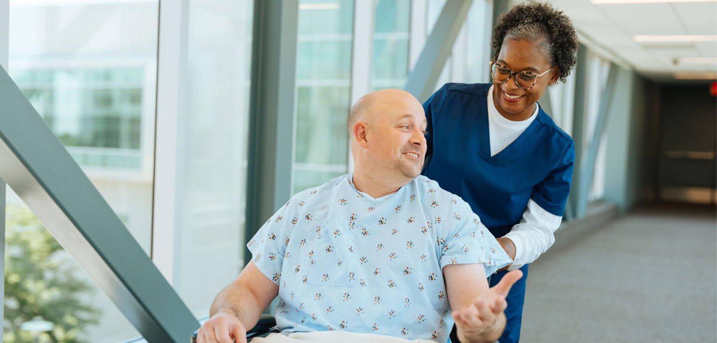 Patient in a hospital gown being pushed in a wheelchair by a healthcare worker along a hospital corridor.