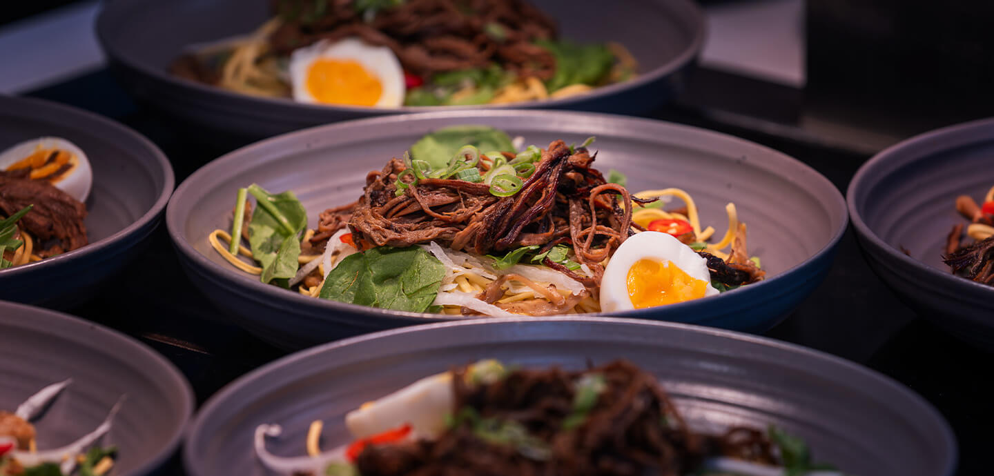 Bowl of noodles topped with shredded meat, vegetables, and a soft-boiled egg, with similar bowls in the background.