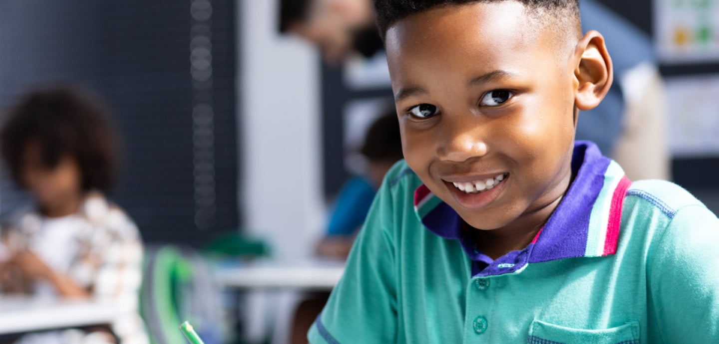 Child sitting at a classroom desk writing with a green pencil, with other students and desks visible in the softly focused background of a bright learning environment.