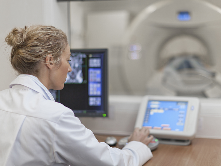 Medical professional in lab coat operates computer while monitoring MRI scan.
