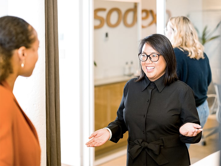 Three people indoors; two facing each other in conversation, one with back turned. Background shows wooden cabinets and a sign reading sodexo.