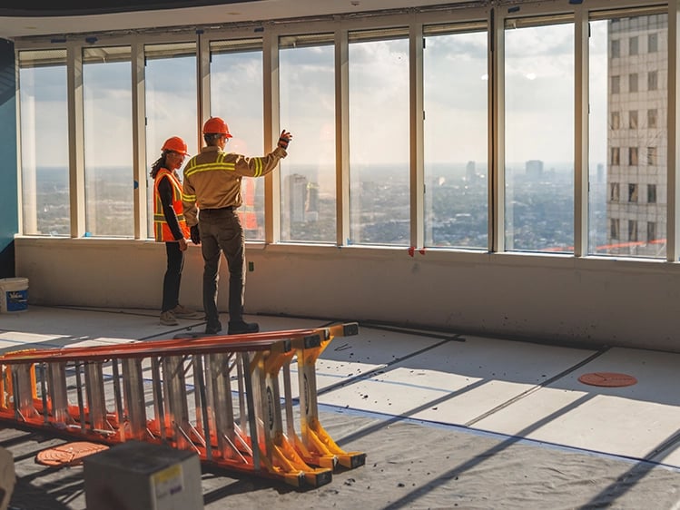 Two construction workers in helmets and vests stand in an unfinished high-rise, looking out large windows at the cityscape; ladders and materials lie on the floor.