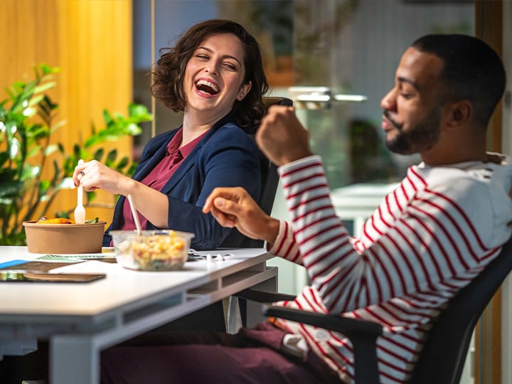 Two people seated at a desk eating lunch from bowls, one gesturing while the other holds a fork, office setting with plants.