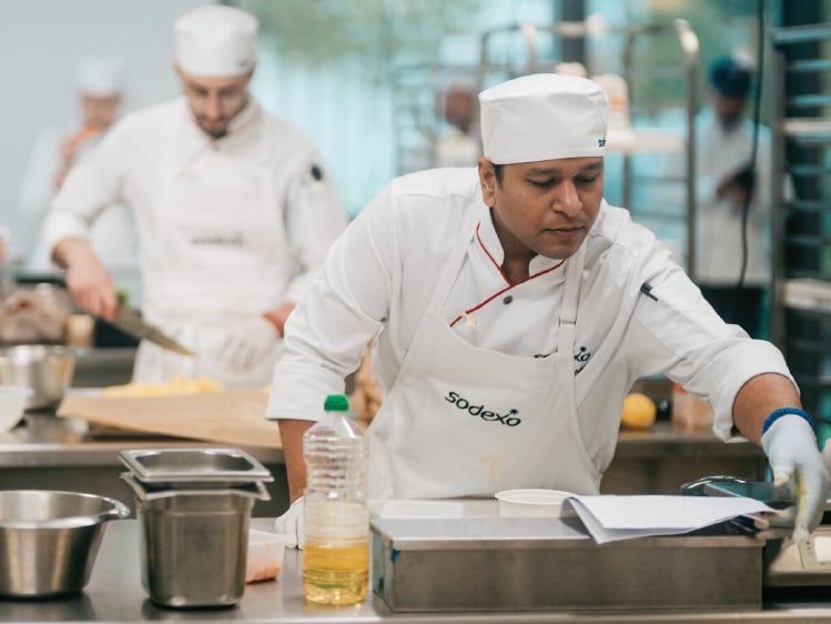 Chef in white uniform preparing food at counter, with another chef working in a professional kitchen behind