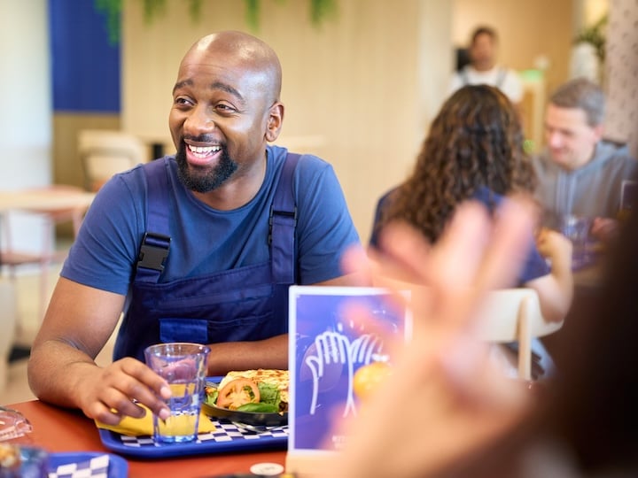 Person in a cafeteria holding a drink while seated at a table with a meal and other diners nearby.