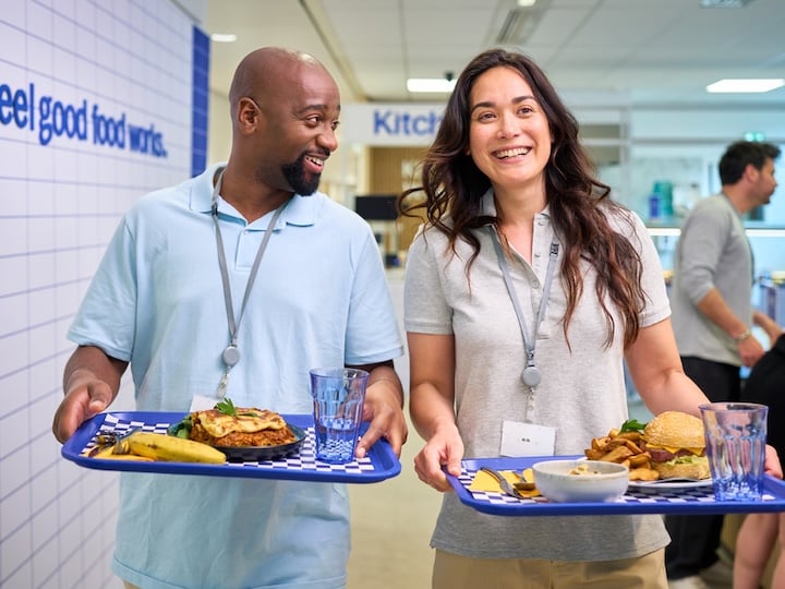 Two cafeteria workers carrying trays of food and drinks while walking through a dining area.