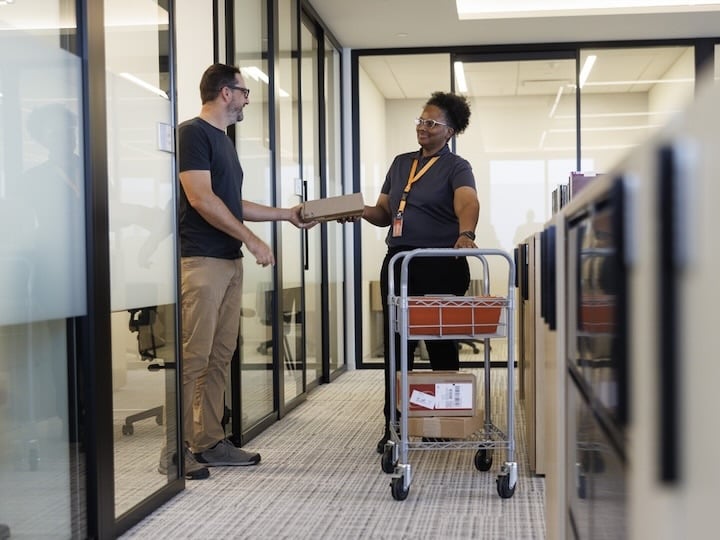 Office worker receiving a package from a delivery cart in a modern office hallway.