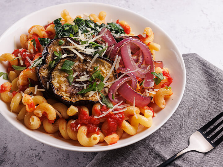 Image of a bowl of cavatappi pasta with tomato sauce, grilled eggplant, sautéed spinach, and red onion, topped with Parmesan and herbs, served with a fork on a gray cloth napkin