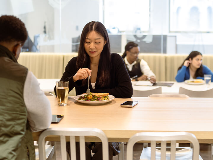 mage of a group of people sitting at a table in what appears to be a modern cafeteria or dining area. In the foreground, a person is enjoying a meal that includes vegetables and possibly meat, accompanied by a glass of beverage and a smartphone on the table.