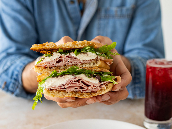 Person holding a stacked deli sandwich with meat, cheese, and greens, with a red drink on the side.