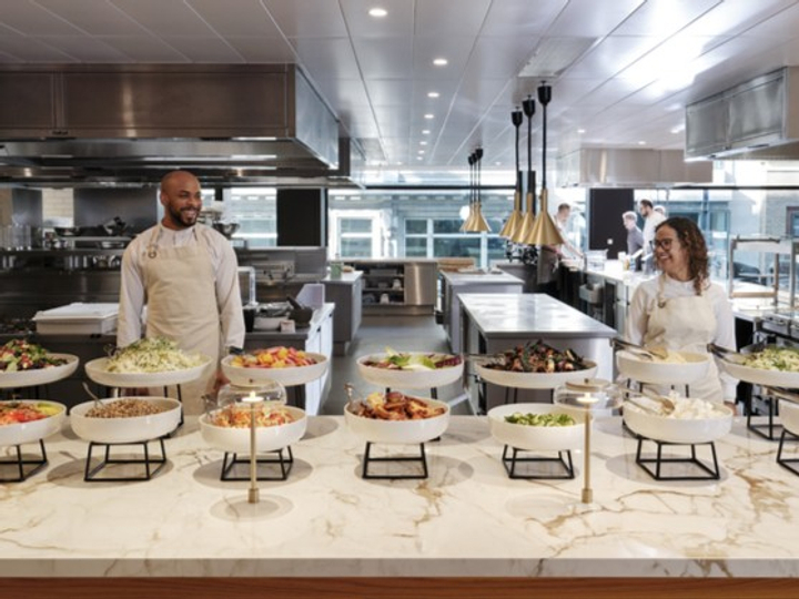 Two chefs behind a buffet counter with assorted salads and dishes in bowls in a modern commercial kitchen.
