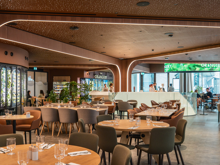 Modern restaurant interior with round tables, upholstered chairs, warm lighting, and open kitchen in background.