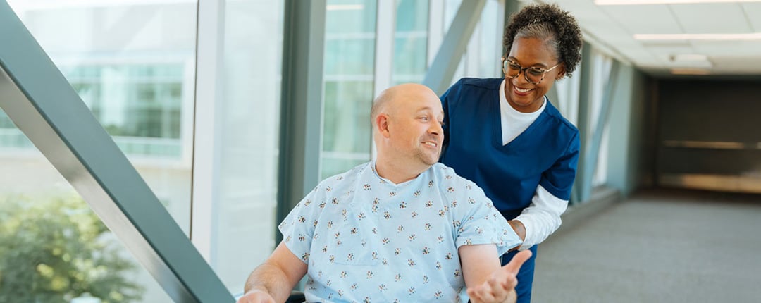 Healthcare professional in blue scrubs assisting a person in a wheelchair in a bright medical corridor.