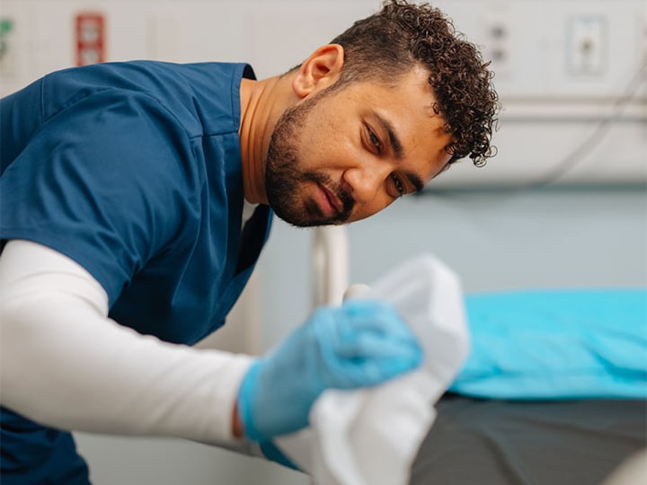 Healthcare worker wiping a hospital bed with disinfectant cloth while wearing gloves in a clinical setting.
