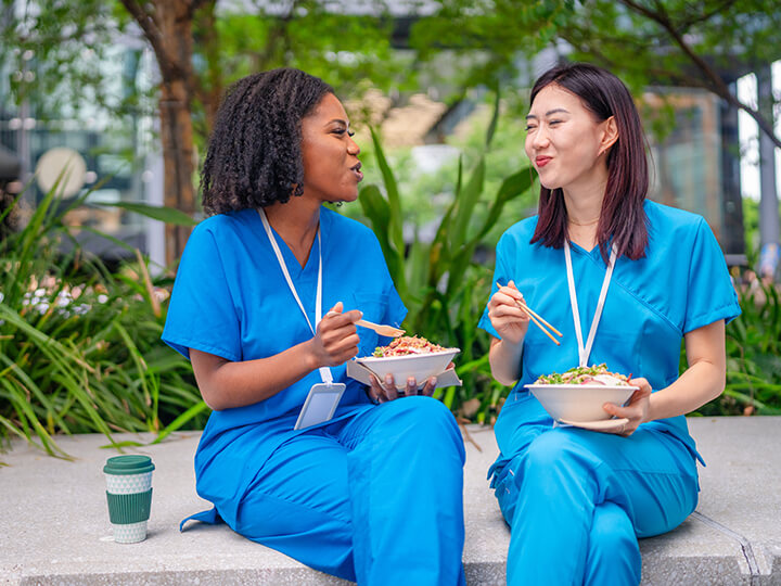 Two healthcare workers in blue scrubs eating lunch outdoors together on a bench.