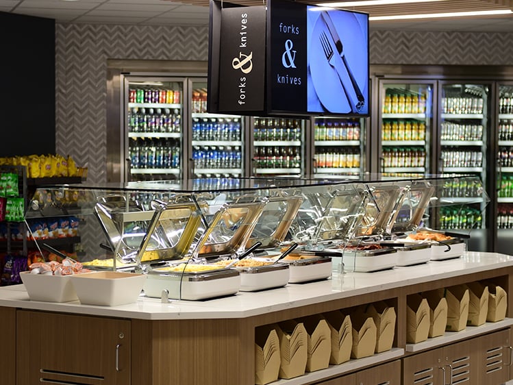 Hot food buffet island in a modern cafeteria with stainless chafing dishes, utensil signage, and drink coolers behind.
