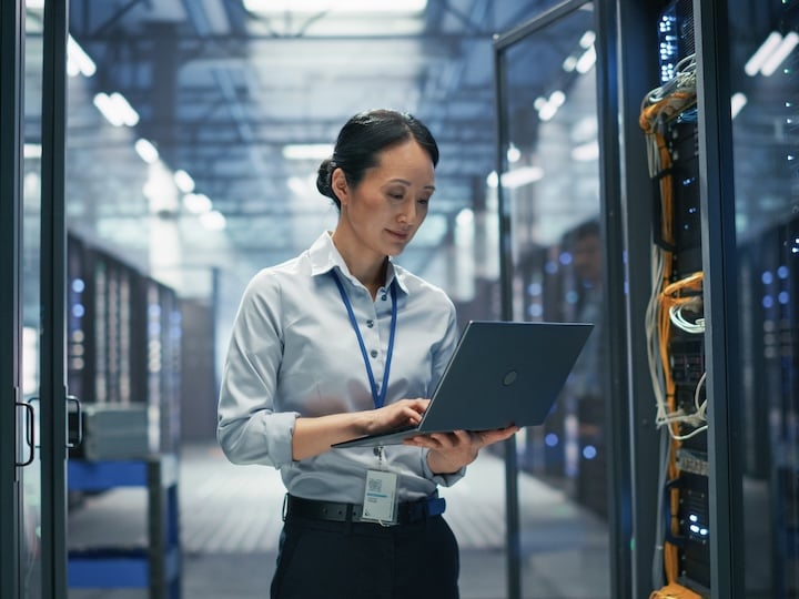 Person using a laptop while monitoring server racks and network equipment in a data center.