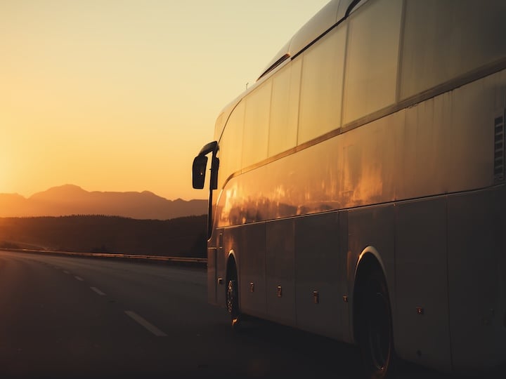 Large coach bus traveling on a highway at sunset with mountains silhouetted in the distance.