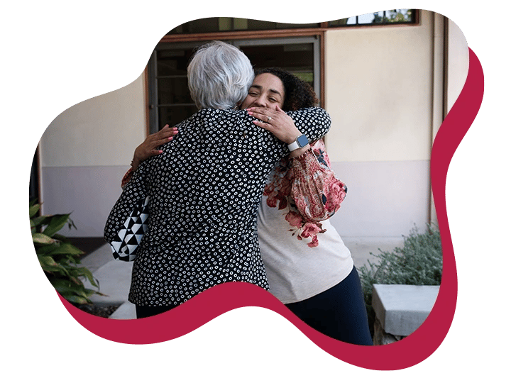 Two people hug warmly outside a building, one in a patterned jacket, the other in a floral blouse.