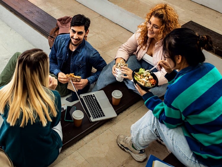 A group of college students sitting on courtyard steps, chatting and sharing lunch together. 