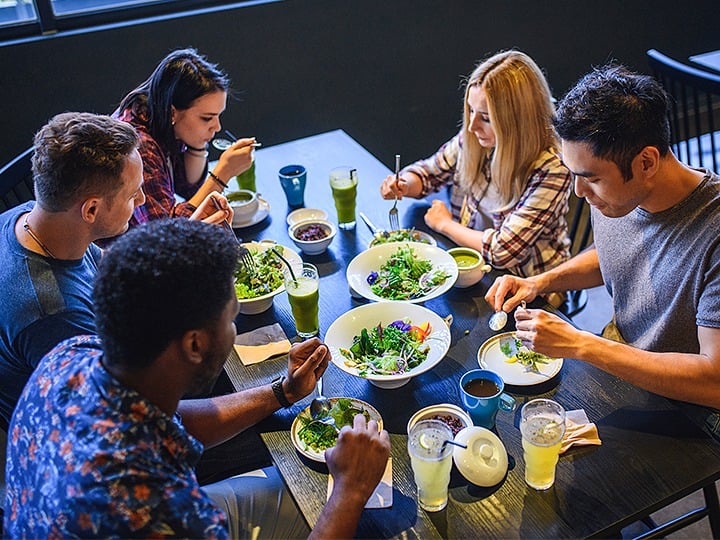 College students gathered around a dining table, serving themselves from a shared salad dish during a casual meal.