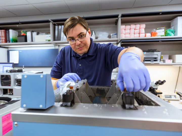 A man in a lab coat and gloves operates a complex machine in a laboratory setting.
