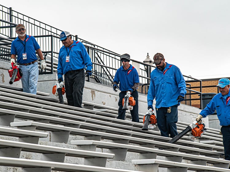 Facilities team members using leaf blowers to clean stadium bleachers, performing outdoor venue maintenance and preparing seating areas for visitors and events.