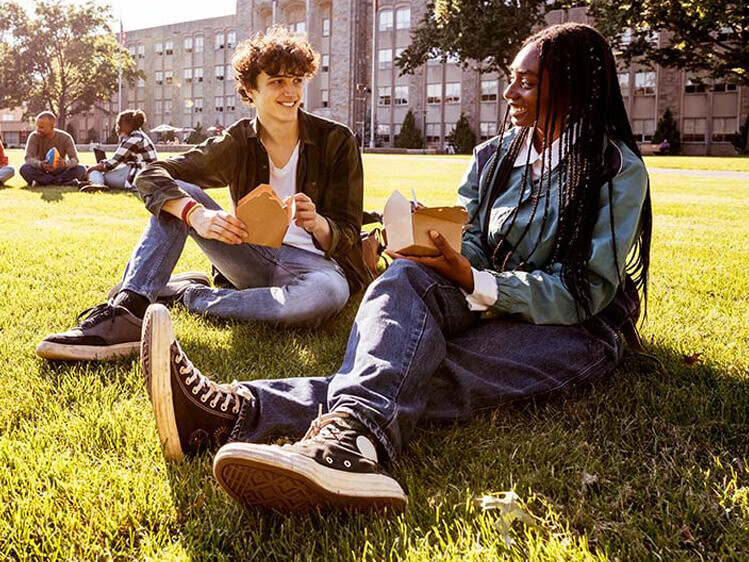 Students sitting on a campus lawn enjoying boxed meals near academic buildings on a sunny day