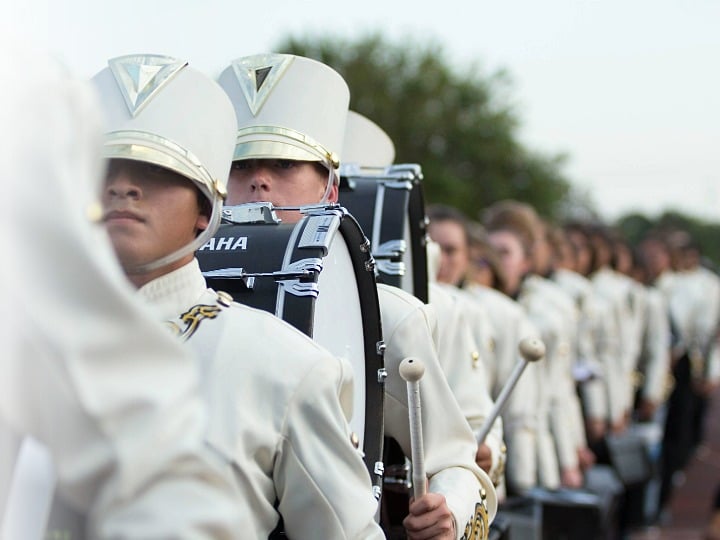 Students in marching band uniforms stand in a straight row holding their instruments.