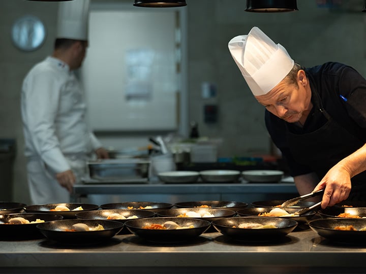 A man wearing a white chef hat, smiling, stands in a kitchen setting with cooking utensils in the background.
