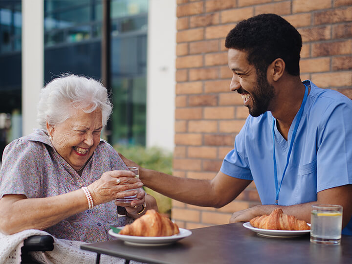 Healthcare worker in blue scrubs sits at an outdoor table with an older adult, sharing a moment over drinks and croissants in front of a brick wall and glass building.