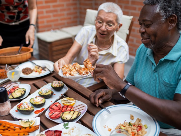 Outdoor table filled with assorted grilled vegetables, stuffed avocados, pasta, and skewers being shared.