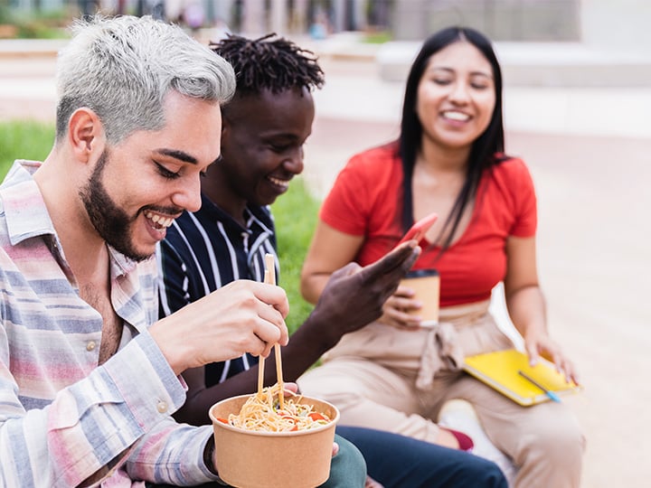 Students sitting outdoors on a campus lawn, eating food from bowls and cups while spending time together in a casual study break setting.