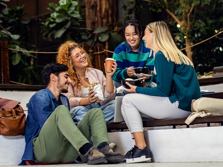 Group of students sitting outdoors on steps, sharing food and drinks while spending time together in a landscaped campus area.