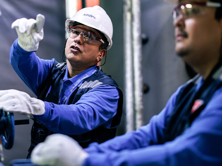 Two workers in blue uniforms and white safety helmets operate industrial equipment, with one pointing while the other adjusts a valve in a technical setting.