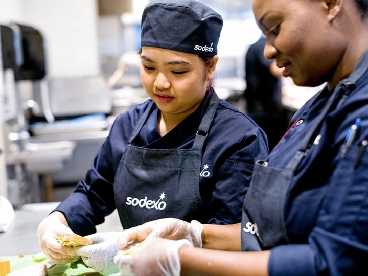 Two women in aprons are preparing food together in a bright kitchen, surrounded by various ingredients and utensils.