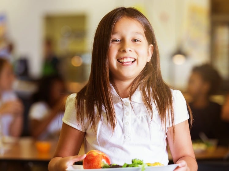A smiling girl in school uniform stands in a cafeteria holding a lunch tray. 