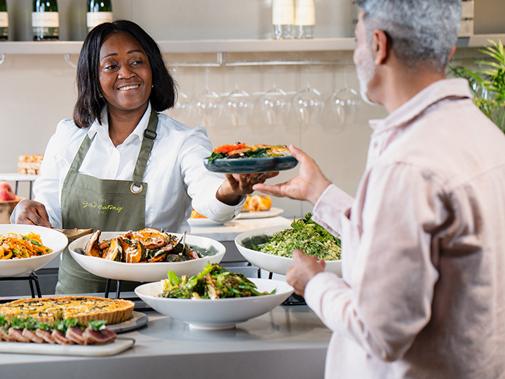 A woman wearing a colorful apron, standing in a kitchen, smiling as she prepares food.