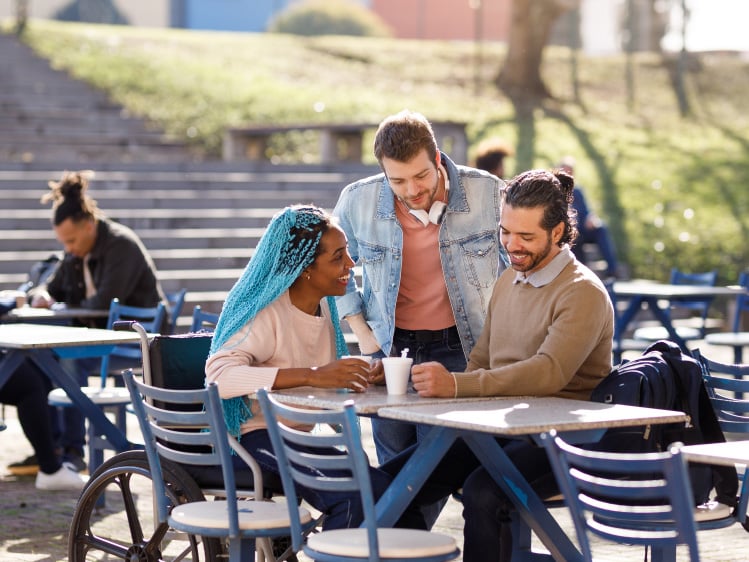 Three people gathered around an outdoor café table on a campus, with one person using a wheelchair, sharing a conversation over drinks in a sunny setting with steps and seating areas in the background.